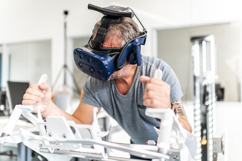 Foto eines Mitarbeiters mit VR-Brille. Er liegt auf dem Icaros-Fitnessgerät und hält sich an Handbefestigungen fest.