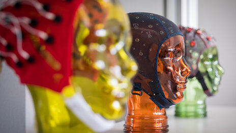 Photo of various EEG masks with stands in the shape of heads on a shelf. The focus is on an orange glass head in the center. __ Various EEG masks with stands in the shape of a face are lined up on a shelf. The focus is on the orange glass head in the middle.