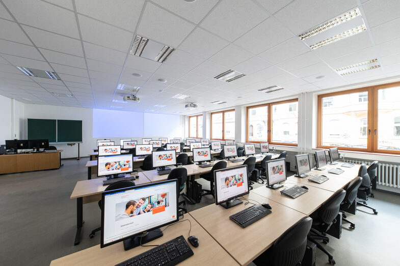 A photo of a computer room. You can see several rows of tables standing one behind the other, with a computer on each table. In the background you can see a blackboard and a projector projecting something onto the wall.