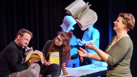 Foto von Schauspieler:innen, die eine Szene im Theaterlabor performen und dabei mit Büchern jonglieren.
