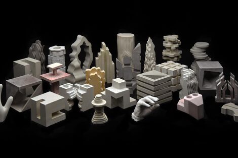 The picture has a narrow horizontal page format. It shows a collection of twenty-seven differently sized, dimensioned and generally differently designed sculptures made of concrete against a black background. The sculptures stand in approximately four rows and are offset in space. Their design appears geometric, free-form or a combination of both. Some of them are colored bright red, grey, black or yellow. At the back are tall, cylindrical or bar-shaped sculptures in terms of volume, but with very individual, sometimes curved and stepped shapes.