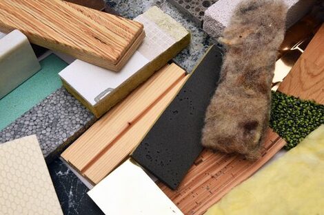 The picture shows a close-up of a disordered collection of different materials in a rectangular shape with the same dimensions and different orientations.
In the foreground of this picture are a multi-layered composite panel made of wood, a piece of composite insulation material, i.e. a piece of rock wool with further layers firmly glued on, a thin strip of black foam, a brown-grey-black, somewhat tattered strip of sheep's wool, a thin, beige-colored strip of Styrodur with a wavy relief on the surface, a white, solid, porous piece of sand-lime brick and a dense, yellow piece of glass wool.
Underneath are other similar pieces of material of this type.