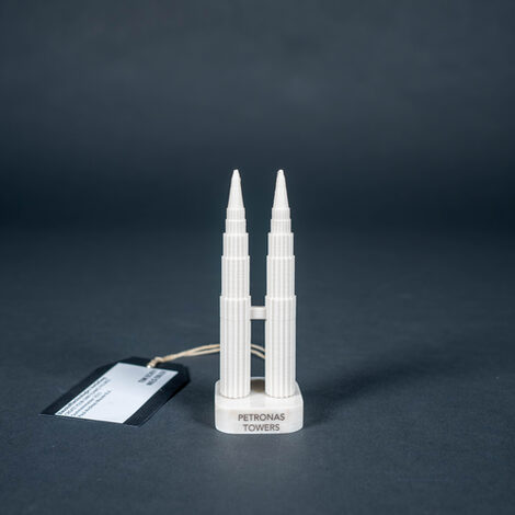 This picture shows a small model of two towers made of white plastic the size of a chess piece. The two towers are the same height, connected by a bridge about halfway up, and they stand on two square marble bases with strongly rounded corners and the name of the building as an inscription. The towers are the Petronas Towers in Malaysia, Indonesia. They look like two pens with vertical grooves, some levels of which are staggered from the tops downwards.