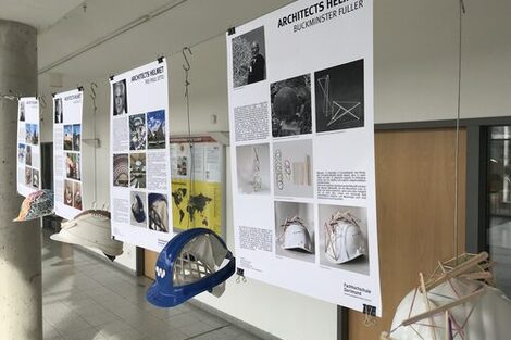 The picture shows four explanatory posters with the corresponding architects' helmets, as they hang in the foyer of the Faculty of Architecture. In the style of Buckminster Fuller or Frei Otto, for example, they are pasted with wooden sticks in a special arrangement or painted in a different color.