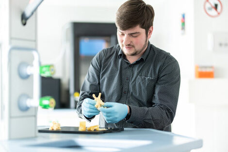 Foto von einem Mitarbeiter mit Handschuhen, der in seinen Händen einen 3D-Ausdruck hält. __ Employee with gloves holds a 3D printout in his hands.