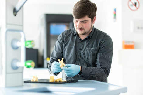 Foto von einem Mitarbeiter mit Handschuhen, der in seinen Händen einen 3D-Ausdruck hält. __ Employee with gloves holds a 3D printout in his hands.