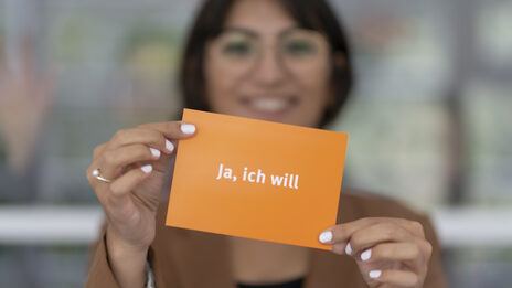 Foto von einer Frau, die eine Postkarte hoch hält. Die Karte ist orange und in weiß mit "Ja, ich will" beschriftet.