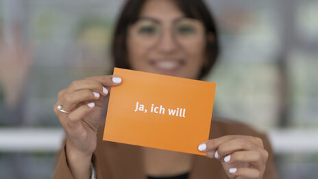 Photo of a woman holding up a postcard. The card is orange and labeled "Yes, I do" in white.