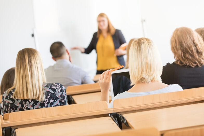 Foto von Hinterköpfen mehrerer Studierender in Sitzreihen im Hörsaal. Vorne ist eine Dozentin unscharf zu erkennen.