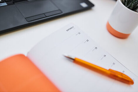 Foto eines aufgeklappten Terminkalenders und eines Kugelschreibers, die auf einem Tisch liegen. Dahinter ein Laptop und ein kleiner Blumentopf im Anschnitt.