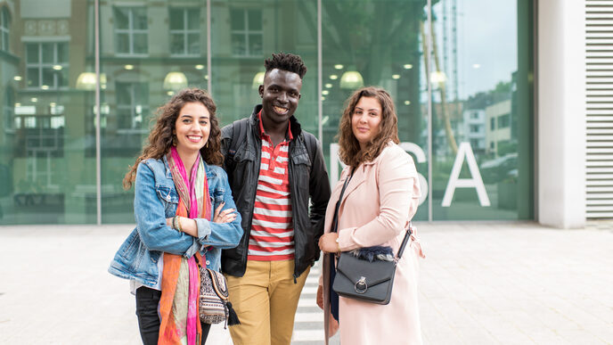 Foto von zwei Studentinnen und einem Studenten, die nebeneinander vor dem Mensagebäude stehen. Sie lachen in die Kamera.__Two female and one male students stand side by side in front of the canteen building. They laugh at the camera.