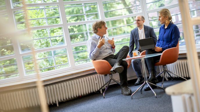 Foto von drei Personen, die vor einem Fenster auf Stühlen sitzen und sich unterhalten. Die Frau rechts hat ein Tablet auf dem Schoß und vor ihnen steht ein Glastisch mit Unterlagen und einer Tasse.