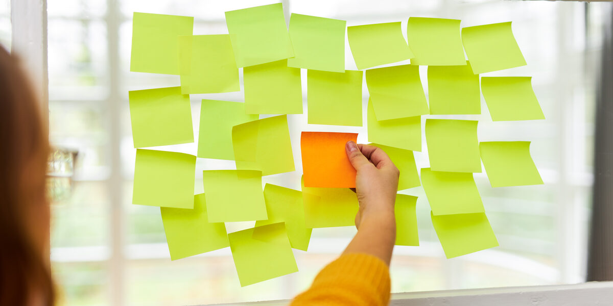 Photo of many yellow Post-its stuck next to and below each other on a window pane. A single orange Post-it is stuck in the middle. A woman stands in front of it and reaches for the orange Post-it.