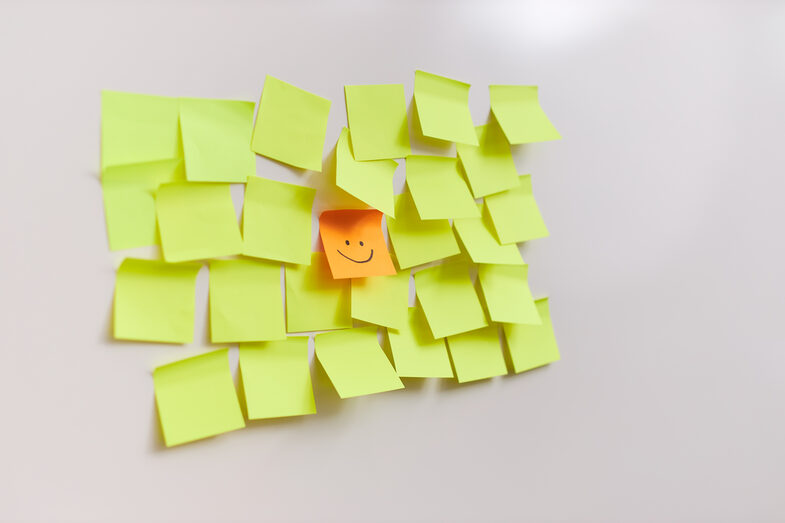 Foto von vielen gelben Post-its, die neben- und untereinander kleben. In der Mitte klebt ein einziges orangefarbenes Post-it mit einem gezeichneten lächelnden Gesicht.