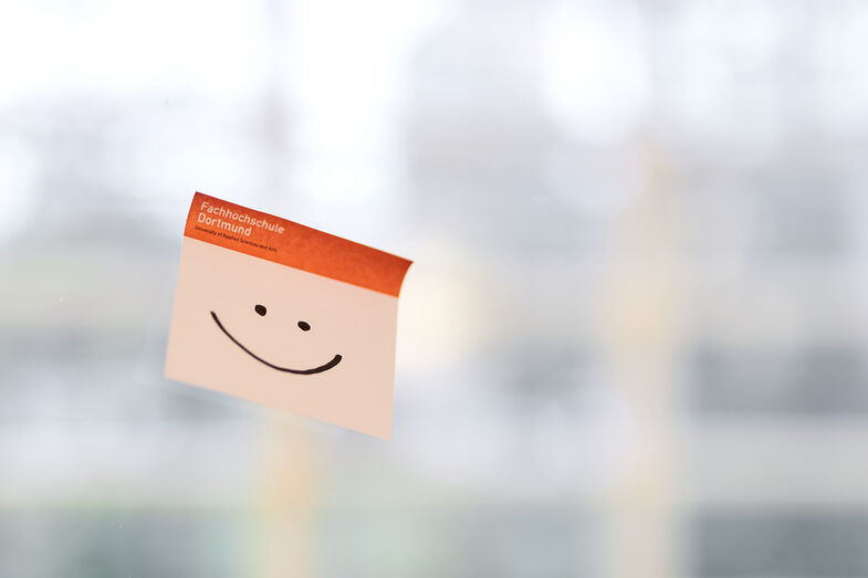 Foto von einem Klebezettel mit FH-Logo, auf dem ein Smiley gemalt ist. Der Zettel klebt an einer Glaswand.