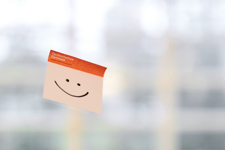 Photo of a sticky note with the FH logo and a smiley face painted on it. The note is stuck to a glass wall.