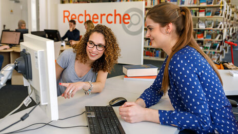 Foto einer Studentin, die einer Mitarbeiterin der Bibliothek etwas mit dem Finger am Monitor zeigt. Im Hintergrund ist unscharf ein Teil der Bibliothek erkennbar sowie Personen an Rechnern.