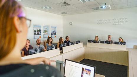 Foto von acht Studierende in einem Seminarraum an PCs. Sie hören einer Frau vorne links im Bild im Anschnitt zu.__Eight students are sitting at PCs in a seminar room listening to a young woman in the front left corner of the picture.