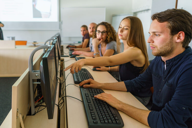 Photo of five students in a row at PCs. One student is showing something to the others on the screen of the student sitting next to her.__Row of five students sitting in a row at five PCs and looking at the screens.__Five students sit in a row at five PCs. One student is showing something to the others on the screen of the student sitting next to her.