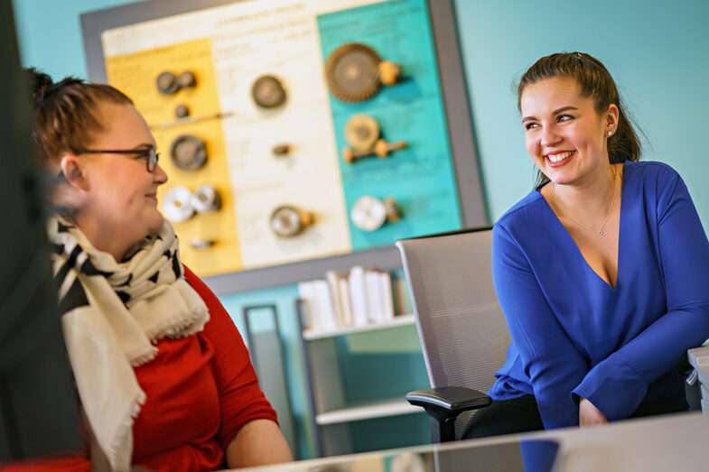 Photo of two library employees sitting next to each other at the reception desk and smiling at each other.