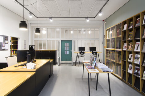 Foto eines Raums in der Bibliothek mit Ausleihtheke, einem Tisch mit Büchern, einem Zeitschriftenregal, einer Vitrine mit historischen Büchern, Schließfächern und zwei Computer-Arbeitsplätzen__Photo of a room in the library featuring a service counter, a table with books, a journal shelf, a display cabinet with historic books, lockers and two computer desks