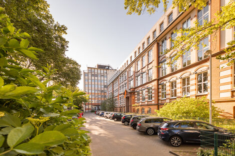 Foto des Altbaus und des Hochhauses an der Sonnenstraße mit Parkplatz davor. Links im Bild im Anschnitt ein blühender Busch.