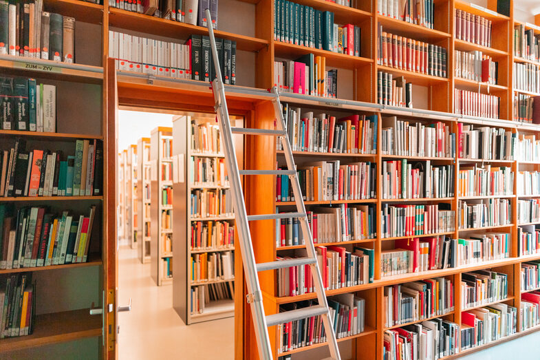 Photo of the library full of books with stairs in front of the shelves and a view through the door to other bookshelves. __