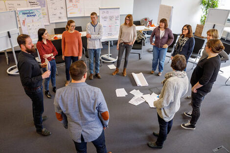 Foto von Mitarbeitenden, die in einem Seminar im Kreis umgeben von Flipcharts stehen. __ In a seminar, employees stand in a circle surrounded by flipcharts.
