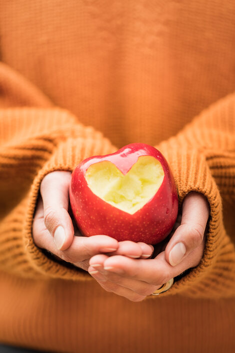 Foto von weiblichen Händen, die gefaltet sind und einen Apfel umschließen, der zur Kamera gehalten wird. In den Apfel ist ein Herz "gebissen". Person trägt orangefarbenen Pullover.