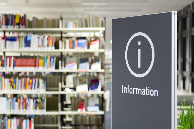 Foto eines Schilds mit der Aufschrift "Information", im Hintergrund ein Bücherregal in der Bibliothek__Photo of a sign reading "Information" with a library book shelf in the background