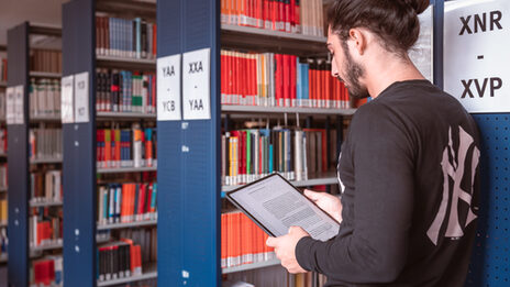 Foot of a young man standing in a library holding a tablet.