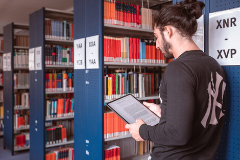 Foot von einem jungen Mann, der in einer Bibliothek steht und in der Hand ein Tablet hält.