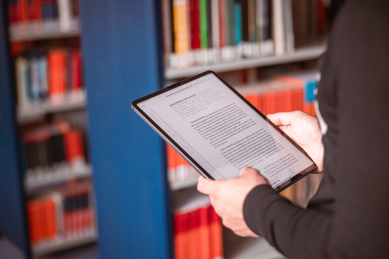 Foto von einem Tablet in der Hand von jemanden, der in einer Bibliothek steht.