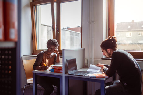 Foto von einer jungen Frau und einem jungen Mann, die mit Büchern und Laptops an zwei Tischen in der Bibliothek lernen.