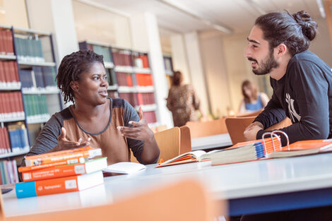 Foto von einer jungen Frau und einem jungen Mann, die an einem Tisch in der Bibliothek sitzen und sich unterhalten. Auf dem Tisch liegen einige Bücher, im Hintergrund sind Bücherregale und zwei weitere lernende Personen zu erkennen.