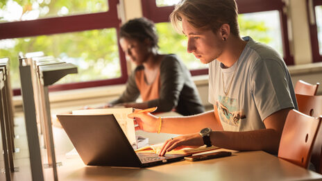 Foto von zwei Studierenden, die mit einem Laptop und Büchern im Lesesaal der Bibliothek lernen.