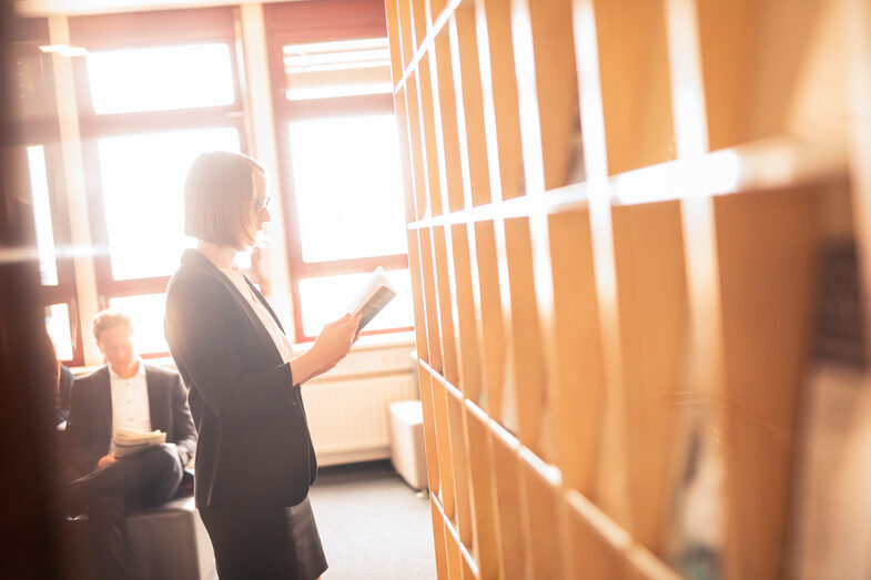 Foto von einer Promovierenden, die in der Bibliothek vor einem Bücherregal steht und in einem Buch liest.