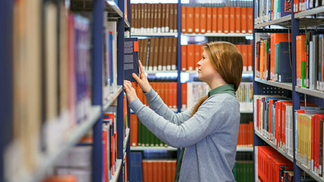 Foto einer Studentin in einer Bücherreihe der Bibliothek. Sie ist zum linken Regal gewandt und holt ein Buch raus.