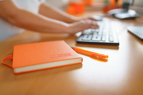 Photo of an FH notebook and FH ballpoint pen lying on a desk. A woman is typing on the keyboard in the background. __ FH notebook and FH ballpoint pen are on the desk, a woman is typing on the keyboard in the background.