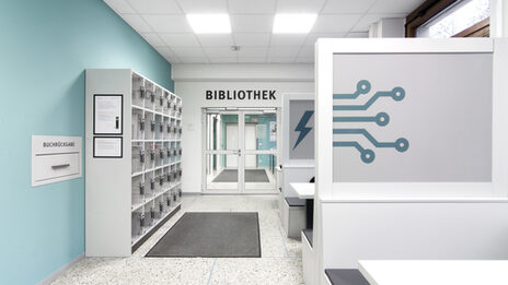 Photo of the hallway in front of the library entrance. It features lockers, a book return box and desks for group study__Photo of the hallway in front of the library entrance. It features lockers, a book return box and desks for group study