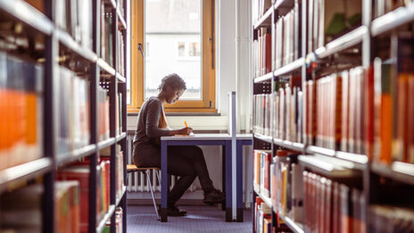 Foto von einer jungen Frau, die an einem Tisch in der Bibliothek zwischen Bücherregalen sitzt und sich Notizen macht.