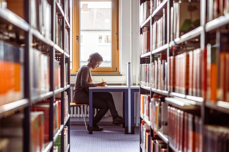 Photo of a young woman sitting at a table in the library between bookshelves and taking notes.