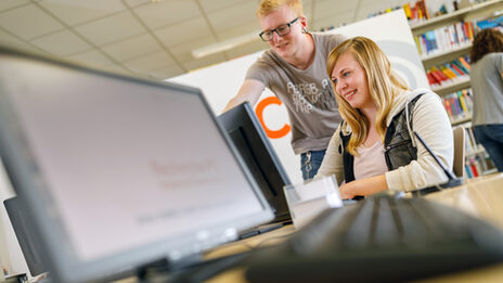 Photo of a male student showing something to a female student on a computer screen at computer workstations in the library __A male student shows something to a female student on a computer screen at computer workstations in the library.