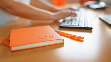 Photo of an FH notebook and FH ballpoint pen lying on a desk. A woman is typing on the keyboard in the background. __ FH notebook and FH ballpoint pen are on the desk, a woman is typing on the keyboard in the background.