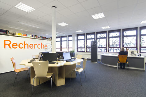 Foto einiger Computer in der Bibliothek, im Hintergrund der Auskunftsplatz, an dem eine Bibliothekarin eine Studierende berät__Photo of computers in the library. In the background, a female librarian is sitting at the reference desk answering a female student's questions