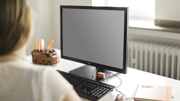 Photo of a woman at her desk, looking at a PC monitor. On the desk is a pencil box and a notebook. __ Woman at work, looking at calculator. On the desk is a pen box and a notebook.