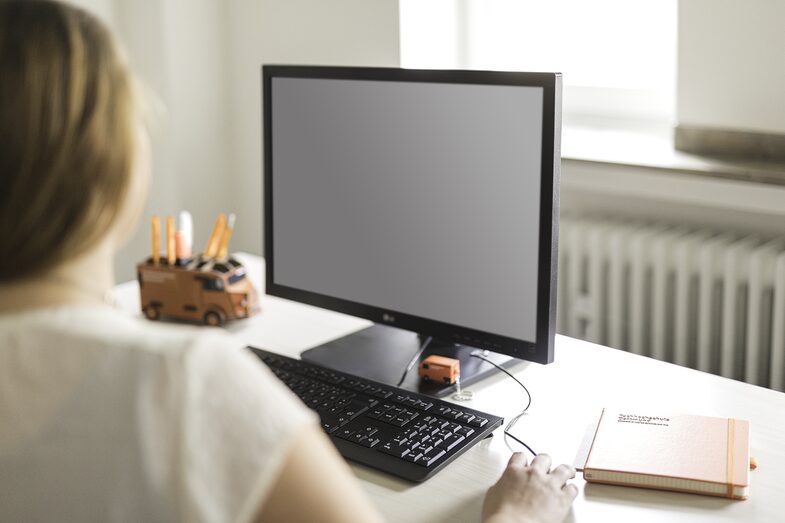 Foto einer Frau am Arbeitsplatz, sie schaut auf einen PC-Monitor. Auf dem Schreibtisch ist eine Stiftebox und ein Notizbuch. __ Woman at work, looking at calculator. On the desk is a pen box and a notebook.