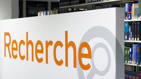 Foto eines Schilds mit der Aufschrift "Recherche", im Hintergrund ein Bücherregal in der Bibliothek__Photo of a sign reading "Recherche" (German for "research") with a library book shelf in the background