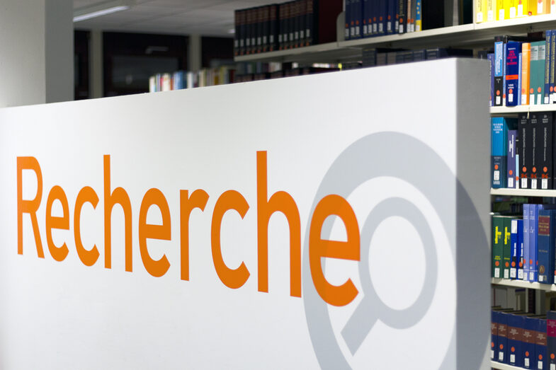 Foto eines Schilds mit der Aufschrift "Recherche", im Hintergrund ein Bücherregal in der Bibliothek__Photo of a sign reading "Recherche" (German for "research") with a library book shelf in the background