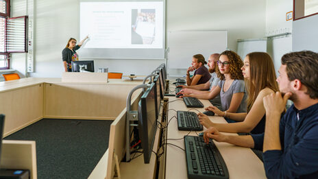 Photo of five students are sitting in a room in a row of tables at PCs, a woman is pointing to something on a projector screen to explain. A woman is pointing to something on a projector screen to explain.__Five students are sitting in a room in a row of tables at PCs, a woman is pointing to something on a projector screen to explain.